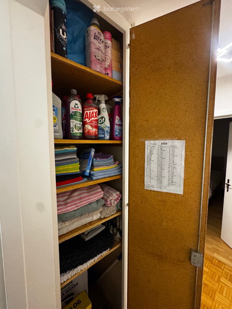 Open wooden shelf unit filled with cleaning supplies and neatly stacked towels. The shelf has multiple levels and is attached to a white wall.
