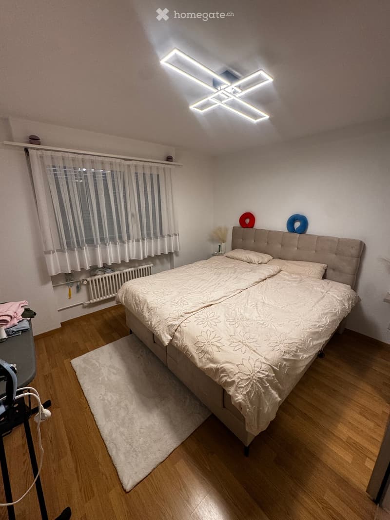 A well-lit bedroom with a large bed, white bedding, a headboard, and a nightstand. The room features wooden flooring, a white curtain, and a radiator under the window.