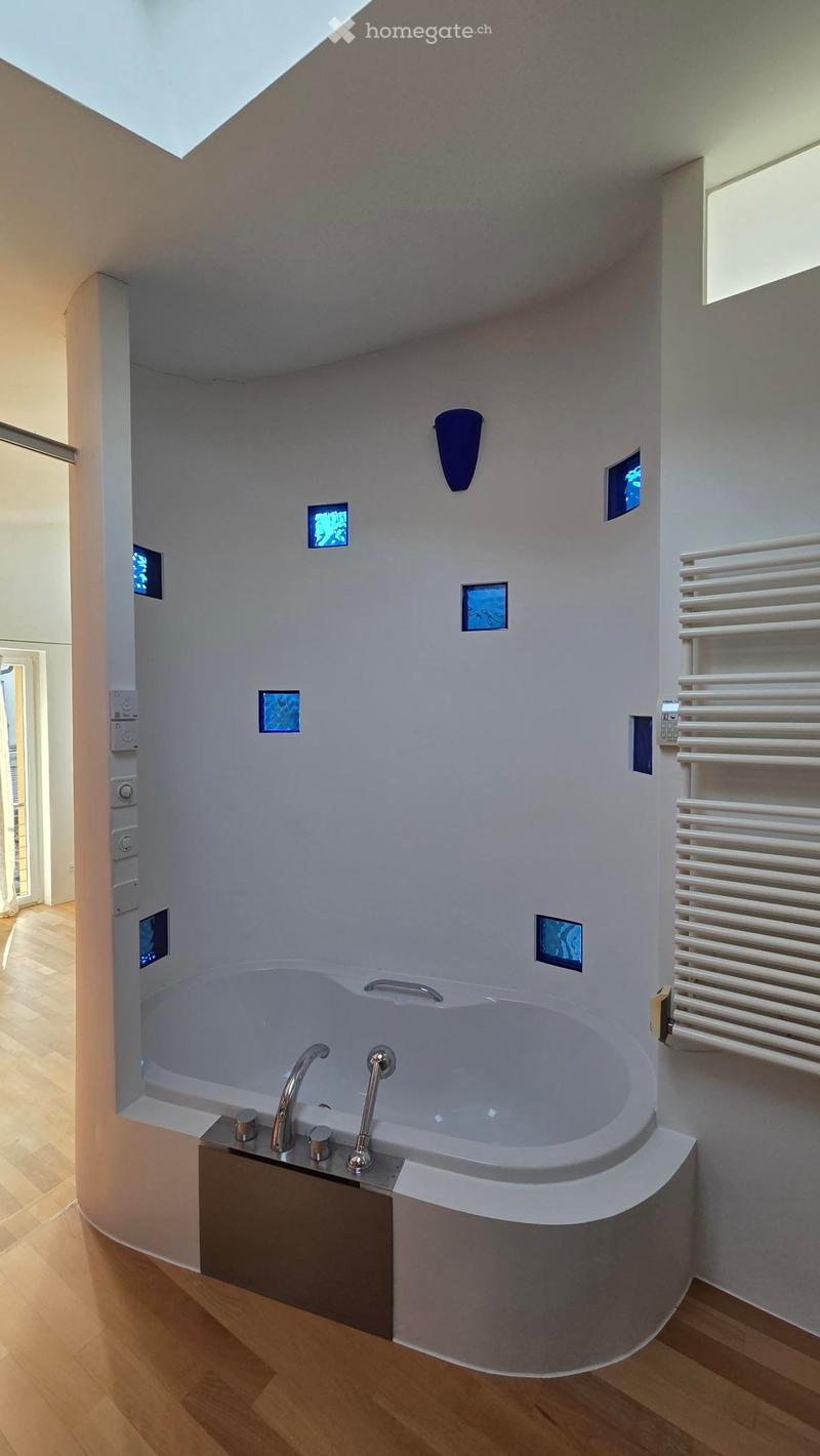 Circular white bathtub, modern faucets, white curved walls, silver handles, blue glass insets, white tiles, wooden floor