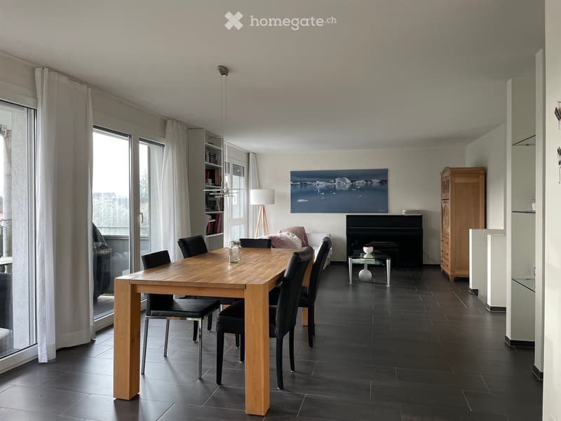 Modern dining area with a wooden dining table, black chairs, a sofa, a piano, and a large picture frame on the wall. The room features large windows with curtains and a floor lamp.