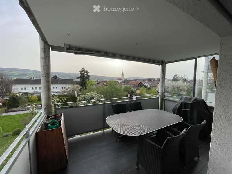 Balcony with a round metal table, wicker chairs, a covered grill, and potted plants, offering a scenic view of a town and surrounding landscape.