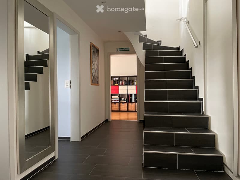 Modern hallway with dark tiled floor, black staircase with metal railing, open doorway leading to another room with bookshelves and storage, framed artwork on wall.