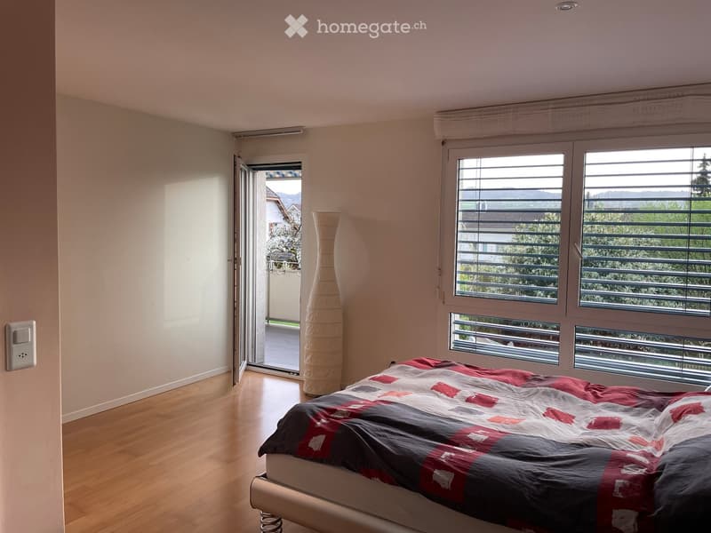 Bright bedroom with wooden flooring, large windows with blinds, a bed with a patterned quilt, and a tall vase near the window.