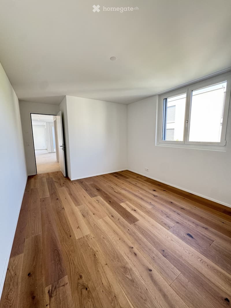 Empty room with light wooden flooring, white walls, and large windows allowing natural light. The room is unfurnished and has an open doorway leading to another space.