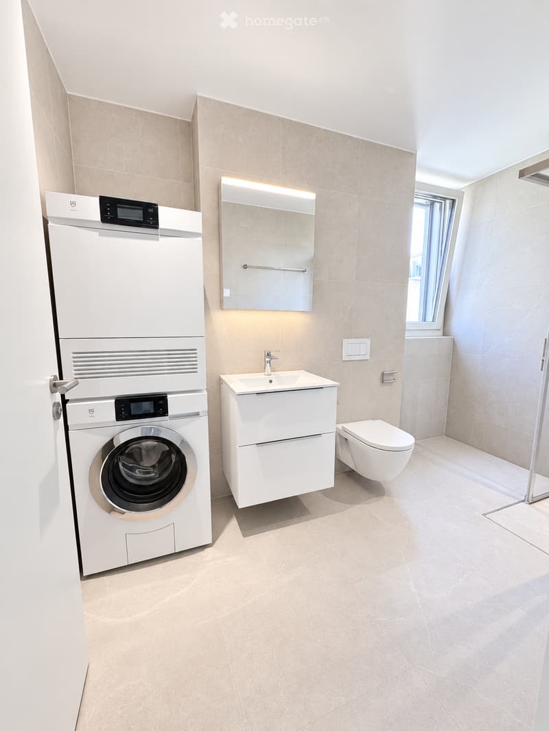 Modern bathroom with beige tiles, white fixtures, a washing machine, a sink with a mirror, and a toilet.