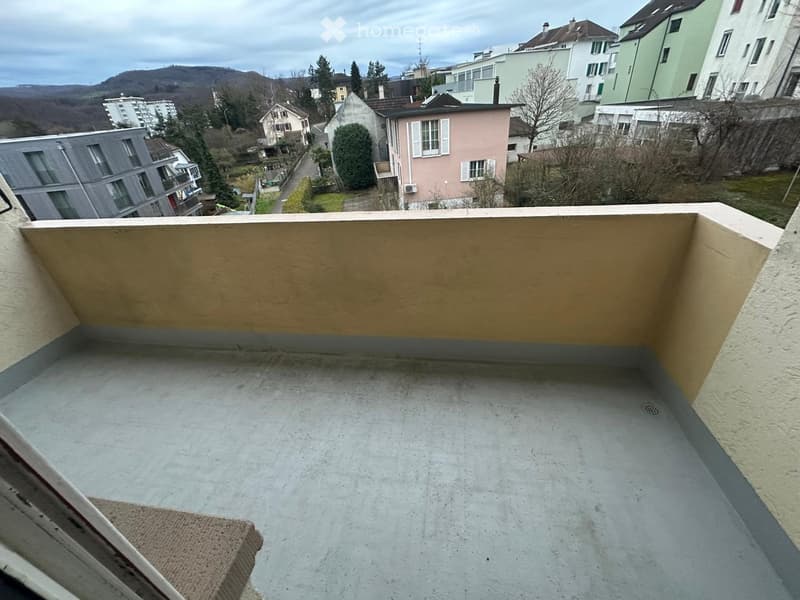 Empty balcony with concrete floor and beige railing, overlooking a residential area with houses and hills in the background.