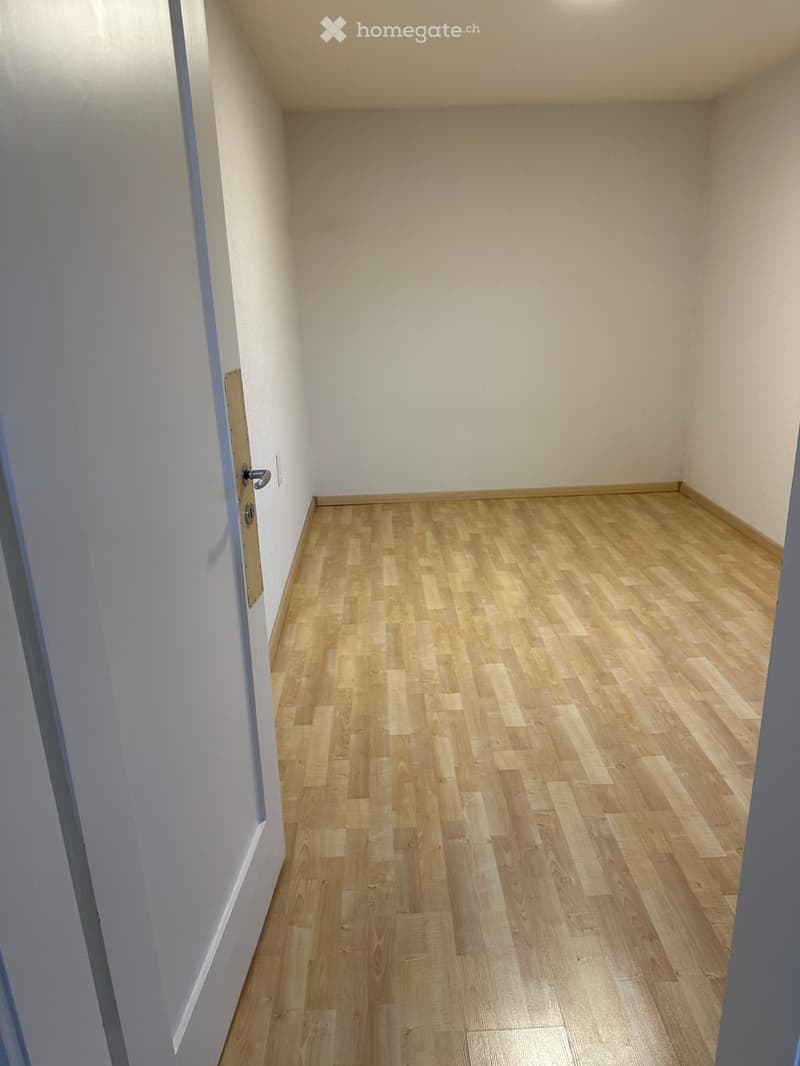 Empty room with light wooden flooring and white walls, featuring a door on the left side.