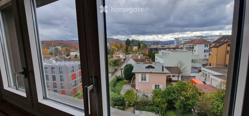 View from a balcony showing a cityscape with residential buildings, trees, and distant hills under a cloudy sky.