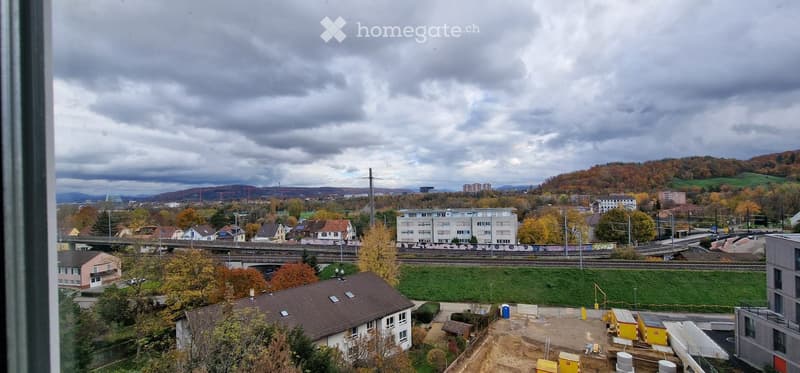 View from a window showing a suburban area with houses, a railway line, and distant hills under a cloudy sky.