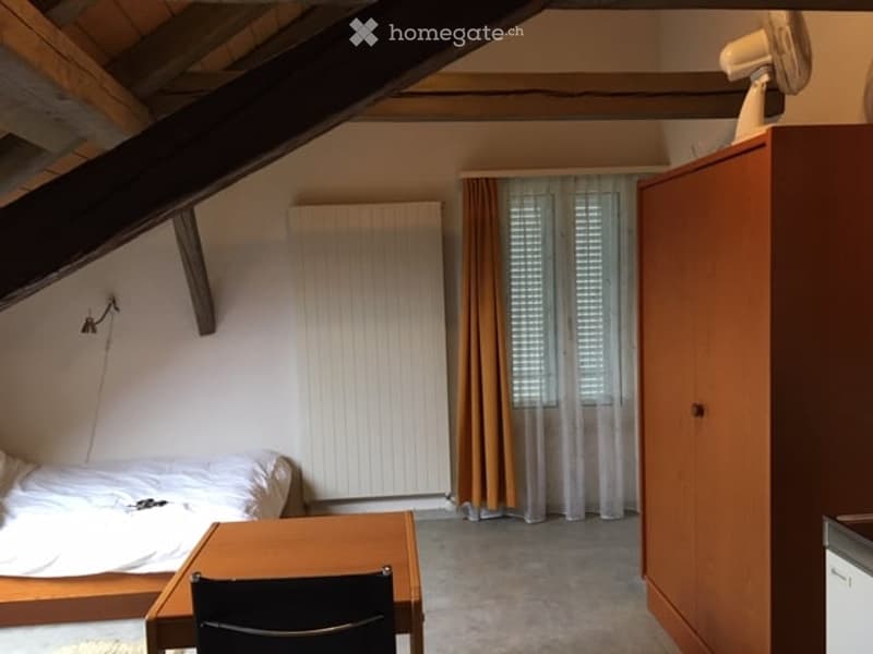 Cozy attic bedroom with slanted wooden ceiling beams, a single bed, a small wooden table with a chair, a wardrobe, and a fan on top of the wardrobe. The room has large windows with blinds and curtains, and a radiator on the wall.