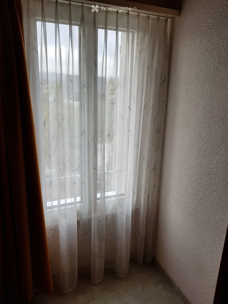 Empty room with white walls, tiled floor, and sheer white curtains on a window, allowing natural light to filter in.