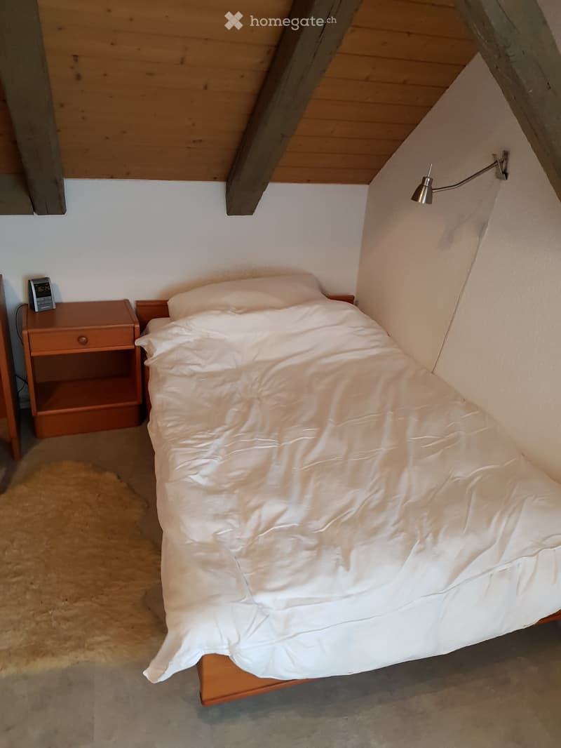 Cozy attic bedroom with a single bed, nightstand, alarm clock, and a wall-mounted lamp. The room features wooden beams and a sloped ceiling.