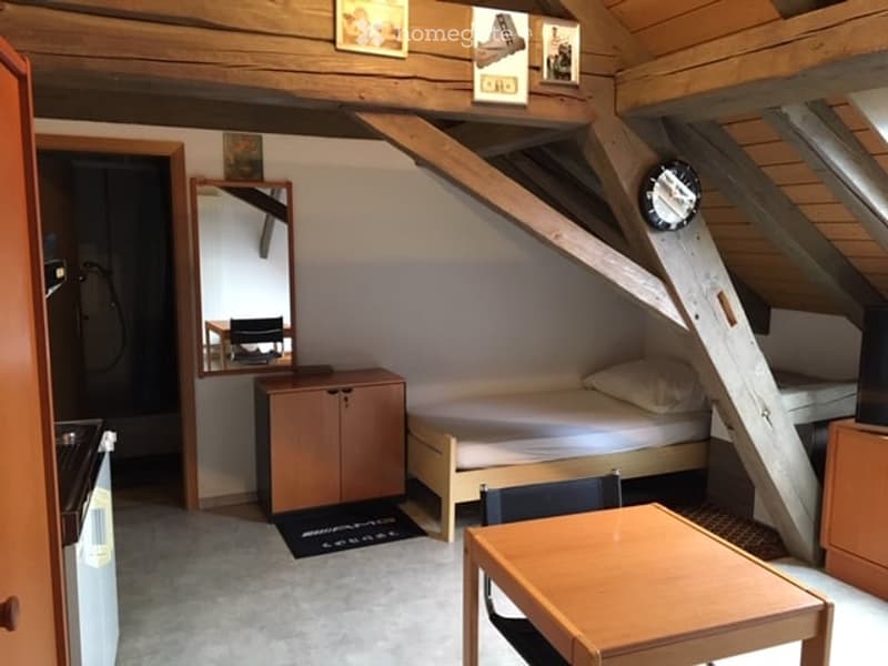 Cozy attic bedroom with wooden beams, a single bed, a small table and chairs, a mirror, and a cabinet.