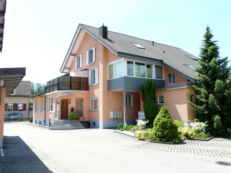 Two-story house with peach-colored exterior, white trim, multiple windows, and a small balcony on the upper floor. The house features a well-maintained garden with trimmed shrubs and trees, and a paved driveway leading to the entrance.