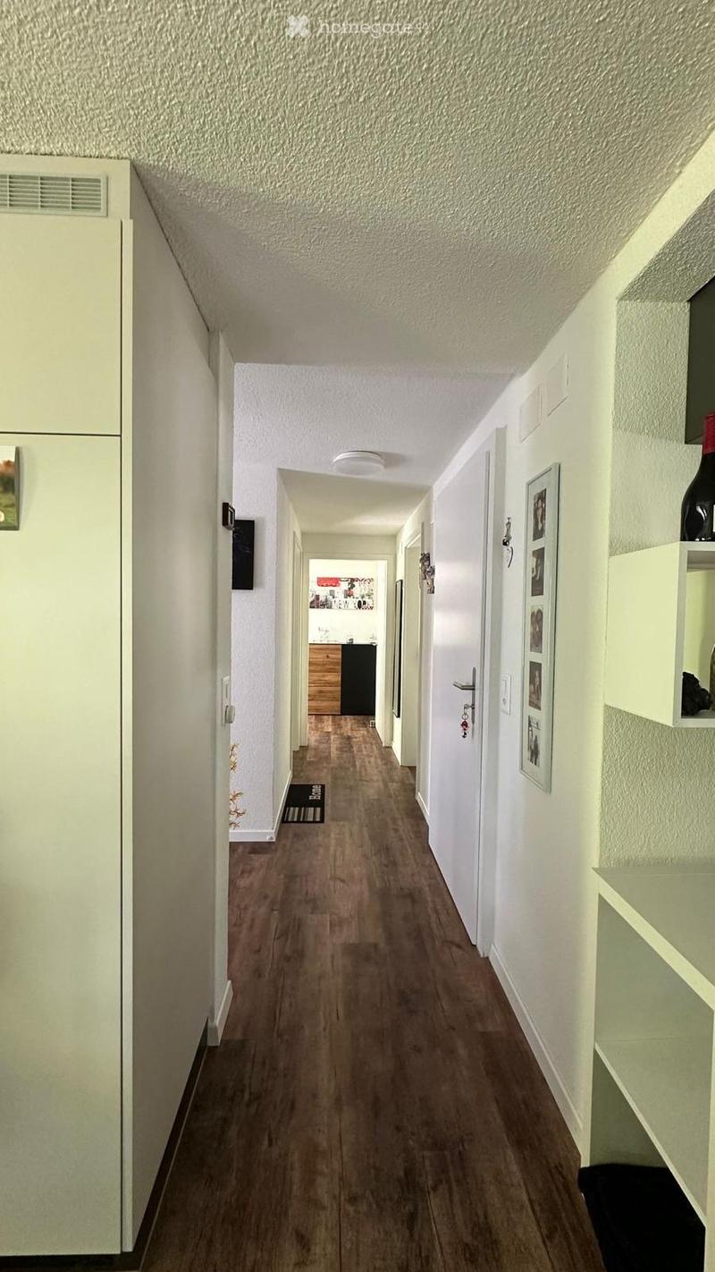 A well-lit hallway with wooden flooring, white walls, and several doors leading to different rooms. The hallway features a small shelf with framed pictures and a bottle on the right side.