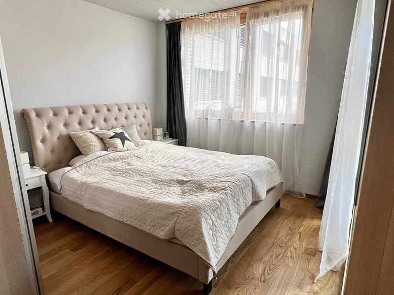 A well-lit bedroom with a large upholstered bed, matching bedding, and a nightstand with a lamp. The room features wooden flooring and large windows with sheer curtains.