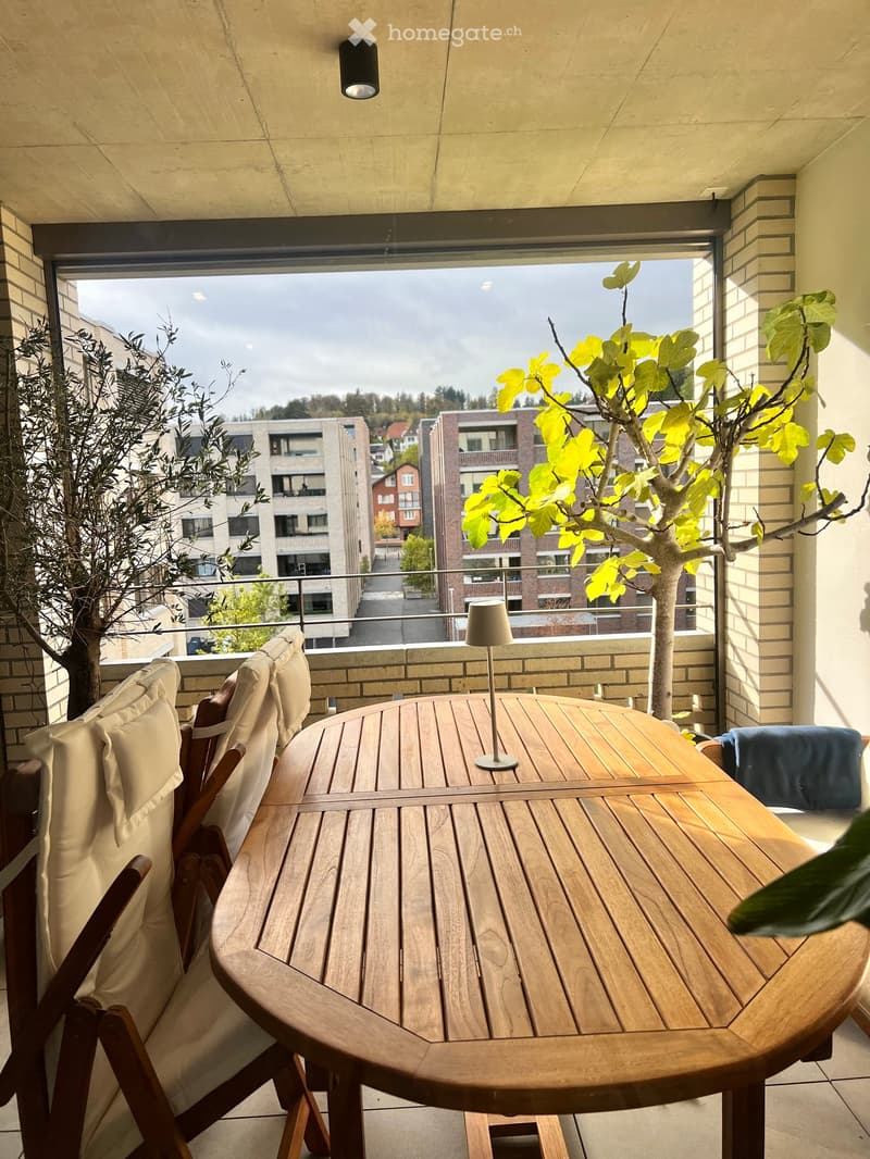 Balcony with wooden table, chairs, and a lamp, overlooking a cityscape with buildings and trees.