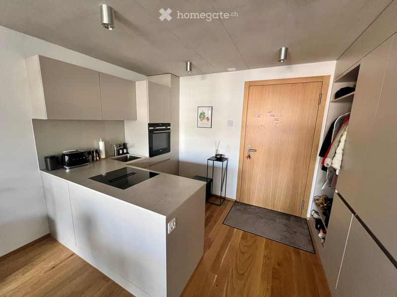 Modern kitchen with light-colored cabinets, induction cooktop, oven, sink, and small table. Wooden floor and white walls.