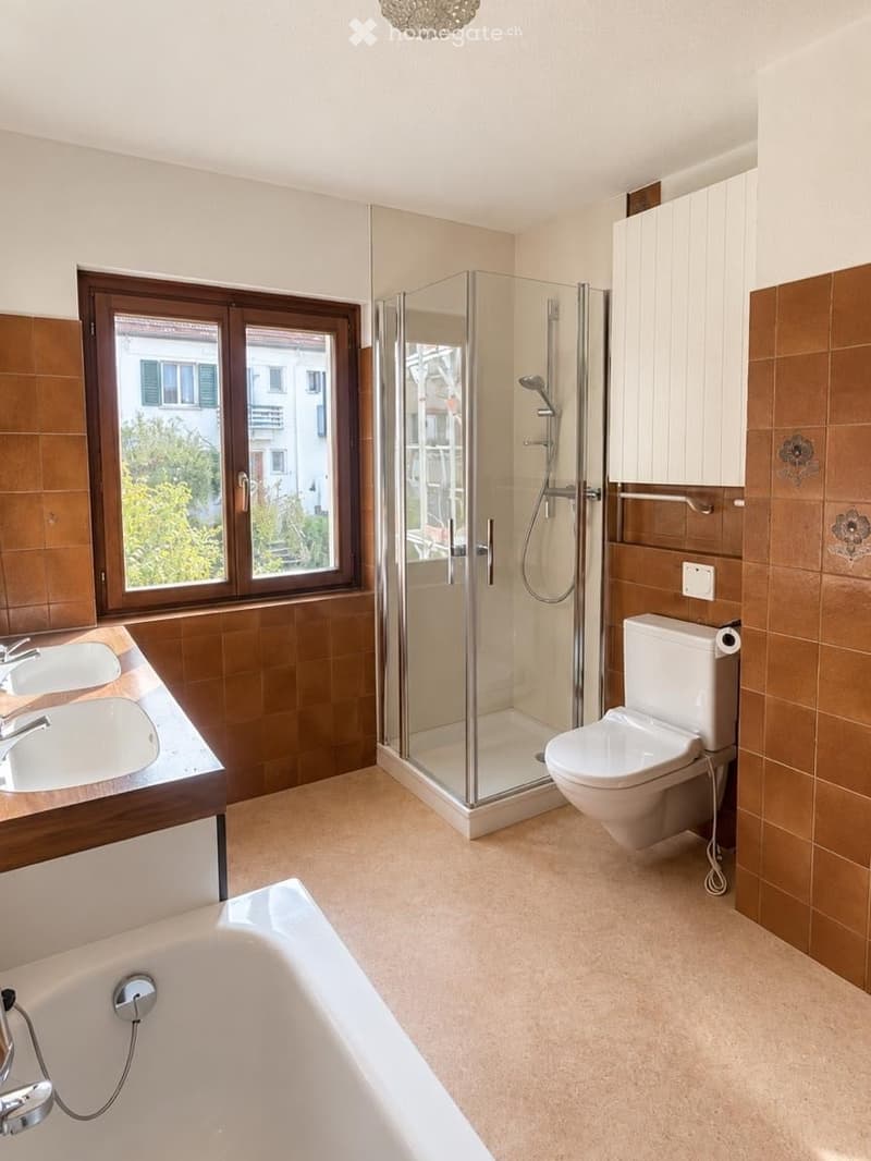 Modern bathroom with a bathtub, toilet, double sink, and a glass-enclosed shower. The room features brown tiled walls and a large window offering a view of the outside.