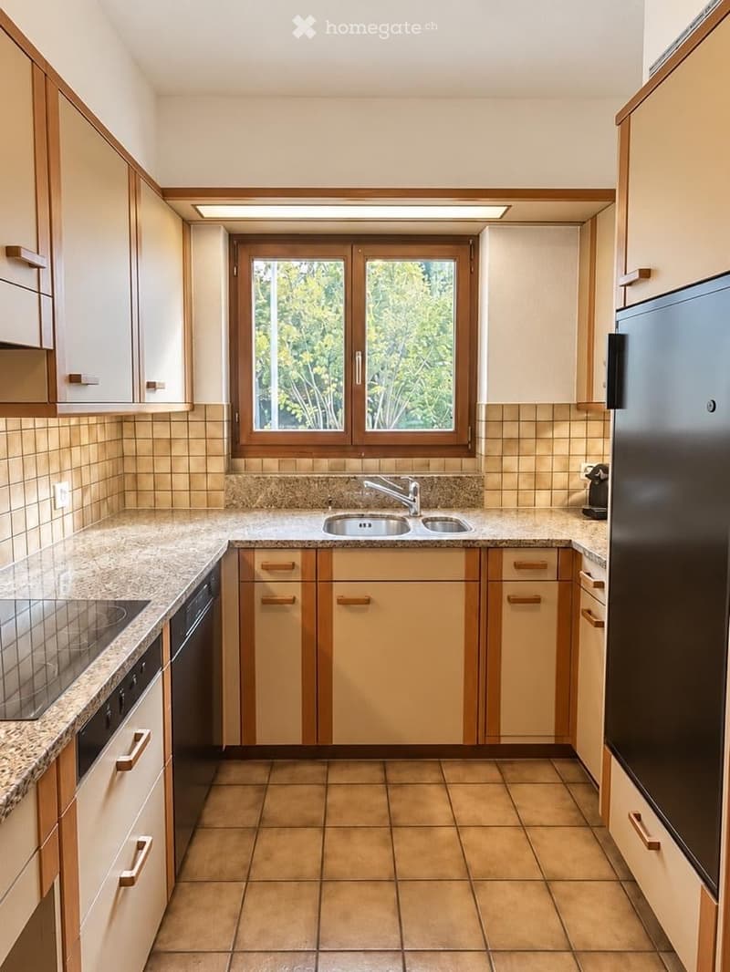 Bright kitchen with light wood cabinets, granite countertops, and a double sink. Features include a refrigerator, dishwasher, and a window overlooking greenery.