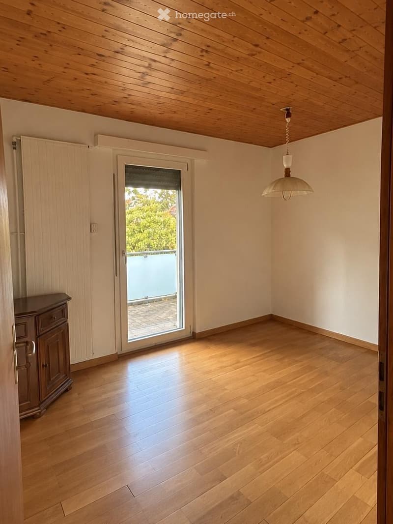 Empty room with wooden flooring, white walls, and a sliding glass door leading to a balcony. Features a wooden cabinet and a hanging light fixture.
