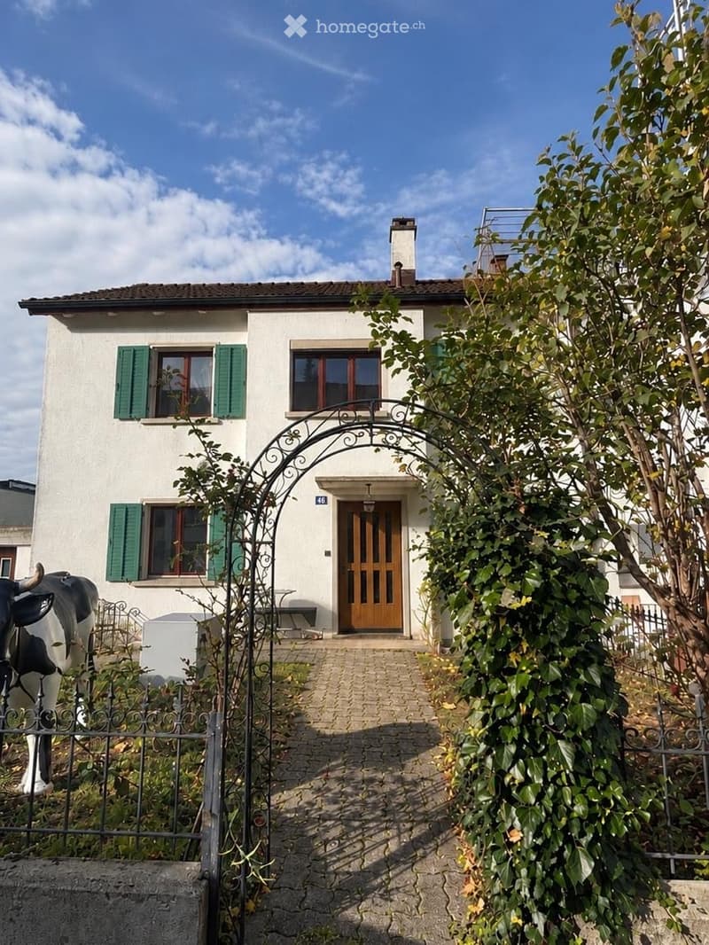Two-story white house with green shutters, wooden front door, and a decorative metal archway leading to the entrance.
