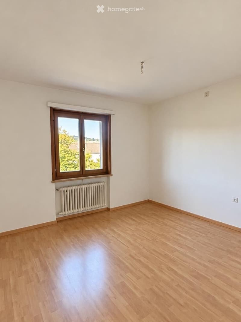 Empty room with wooden flooring, white walls, and a large window with wooden frame. A radiator is placed below the window.