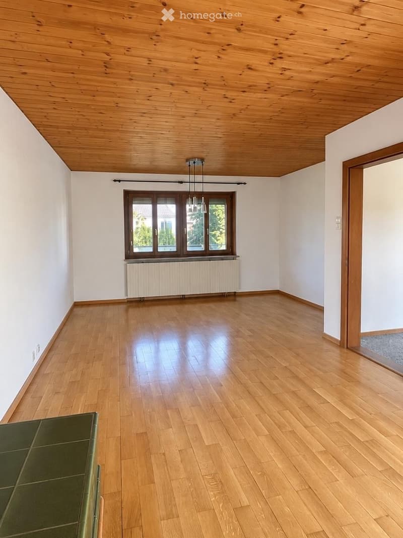 Empty room with wooden flooring, white walls, and a large window with a radiator below. The ceiling is wooden with exposed beams and a hanging light fixture.