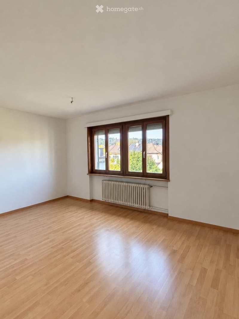 Empty room with wooden flooring, white walls, and a large window with brown frames. A radiator is placed beneath the window.
