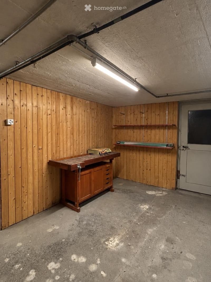 Empty basement or storage room with wooden panel walls, concrete floor, workbench, shelves, and a door.