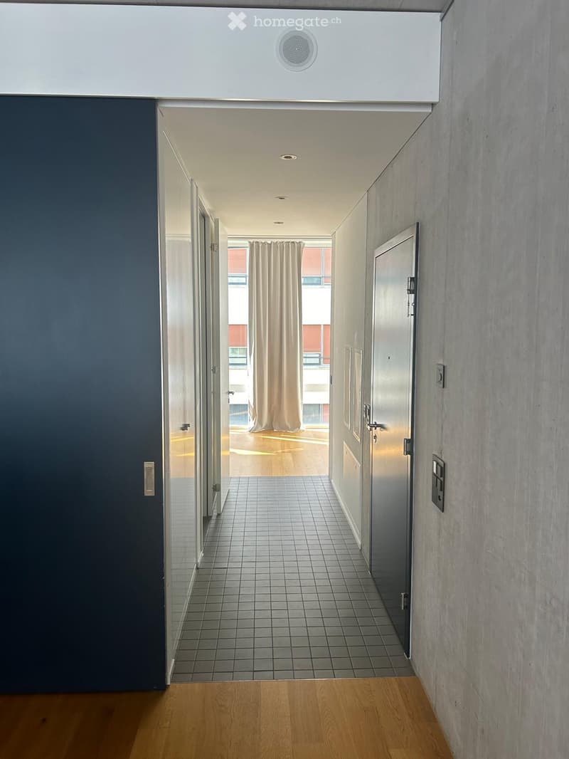 Modern hallway with tiled floor, wooden entrance area, and visible speaker in the ceiling. Doors lead to adjacent spaces.
