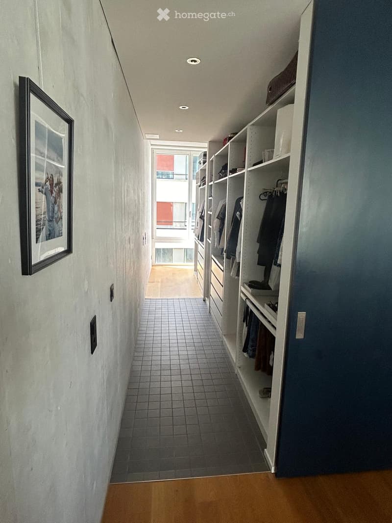 A narrow hallway with tiled floor, built-in closet with hanging clothes, and a framed picture on the wall. The hallway leads to a bright area with a view of a balcony.