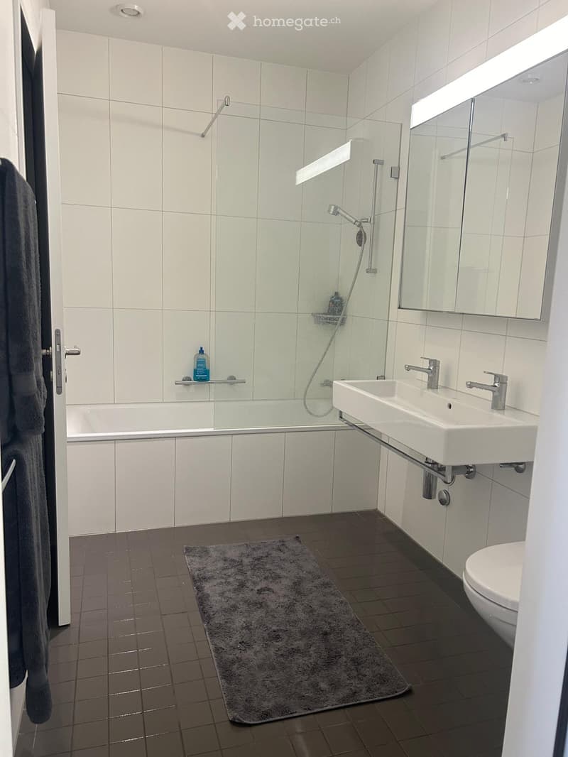 Modern bathroom with white tiles, bathtub, sink, toilet, mirror, and a shower area with glass enclosure. Includes a towel and a bath mat on the floor.
