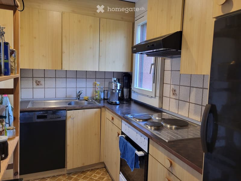 Compact kitchen with light wooden cabinets, stainless steel sink, dishwasher, electric stove, and refrigerator. Countertop has a coffee maker and other small appliances.