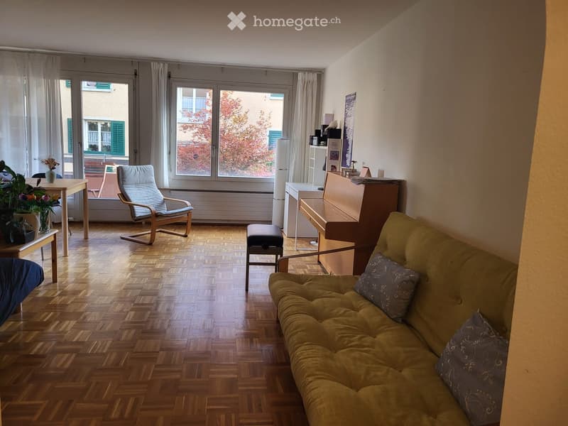 A cozy living room with wooden flooring, large windows, a sofa, a chair, a small table with a plant, a piano, and a stool.