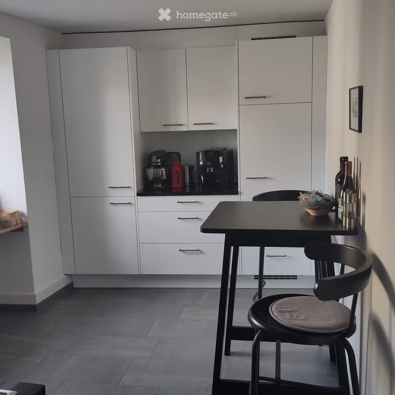 Modern kitchen with white cabinets, a black countertop, a small dining table with a chair, and various appliances including a coffee maker and mixer.