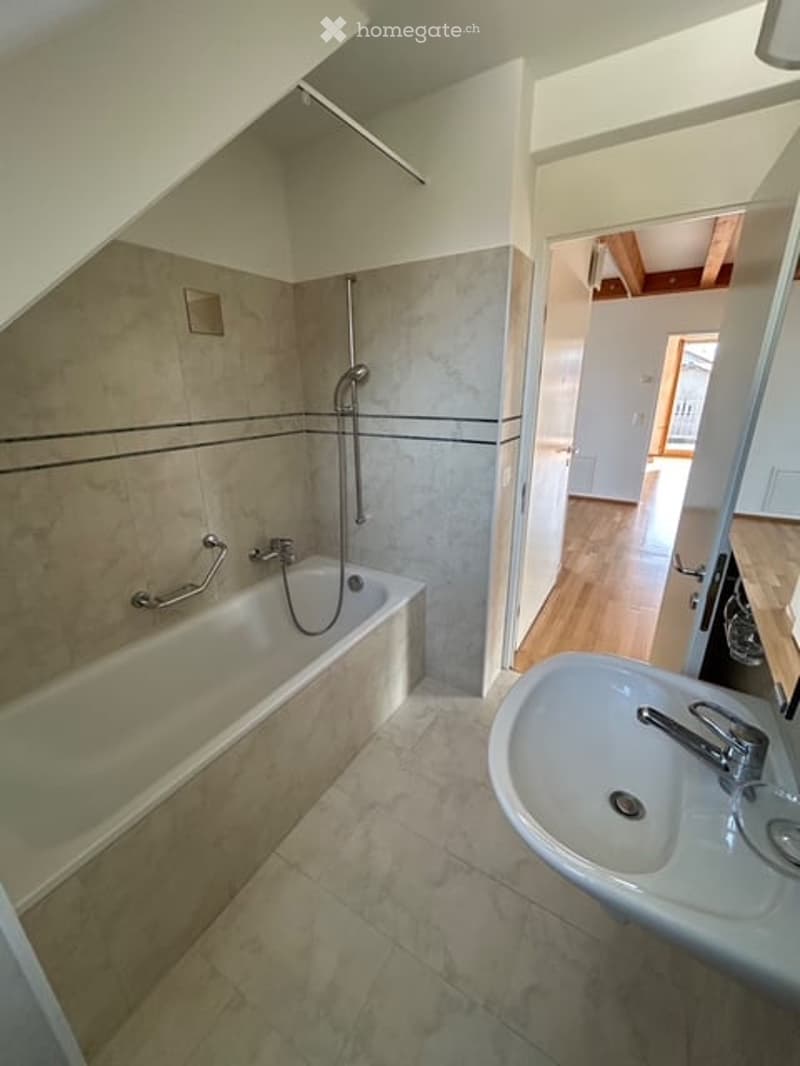 Modern bathroom with a bathtub, sink, and shower hose. Light-colored tiles and wooden flooring are visible in the adjacent room.