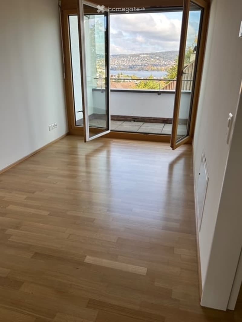 Empty room with wooden flooring, large open sliding doors leading to a balcony with a scenic view of a city and water.