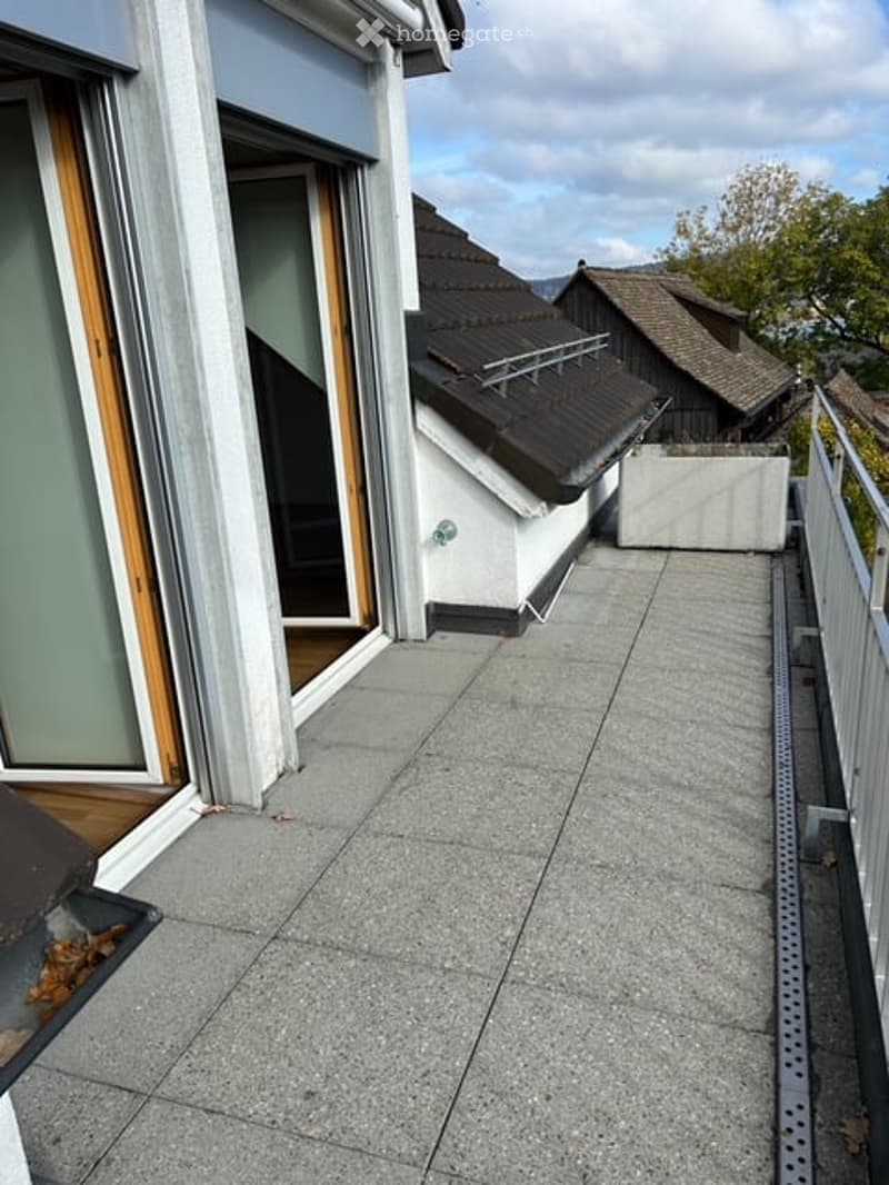 Balcony with grey concrete tiles, metal railing, and a view of neighboring buildings and trees.