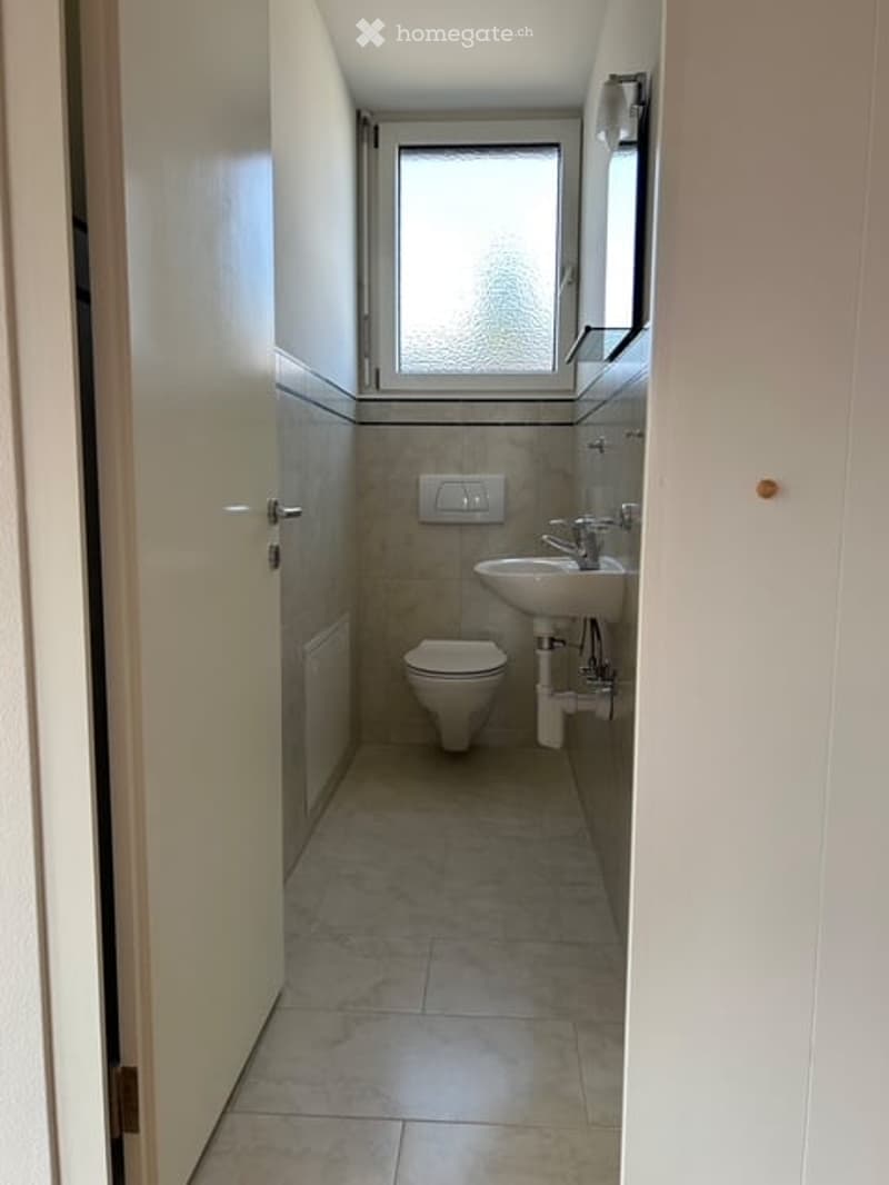 Small, modern bathroom with a toilet, sink, and mirror. The space features tiled walls and floor, with a window providing natural light.