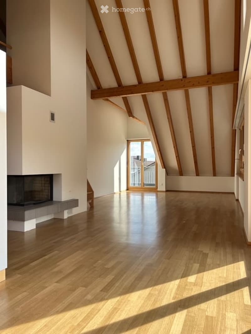 Bright, airy attic space with exposed wooden beams, polished hardwood floor, and a built-in fireplace. Large windows and a glass door lead to a balcony.