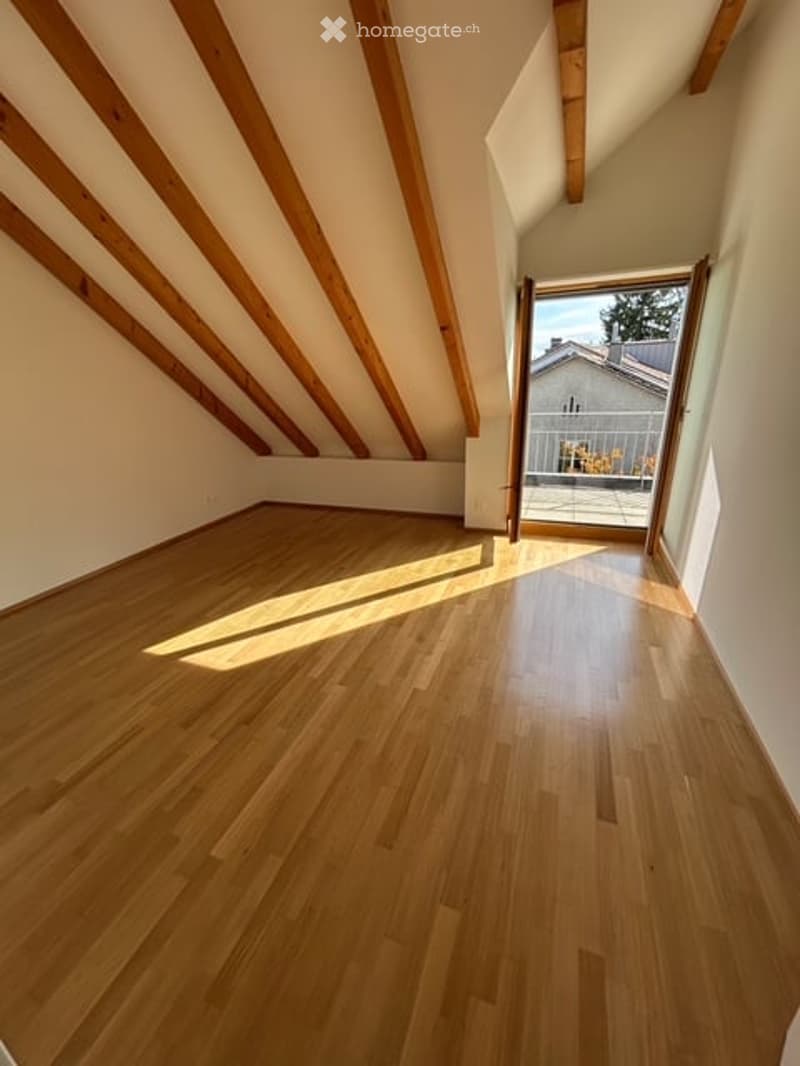 Bright, empty attic room with wooden floors, exposed wooden beams, and a large window opening to a balcony.