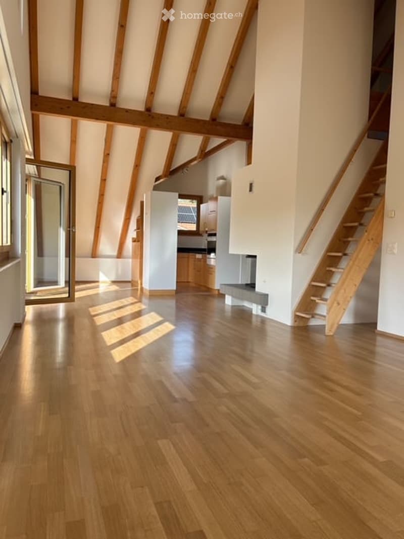 Bright, open loft space with exposed wooden beams, hardwood flooring, and large windows allowing natural light. The room features a staircase leading to an upper level and a partially visible kitchen area in the background.