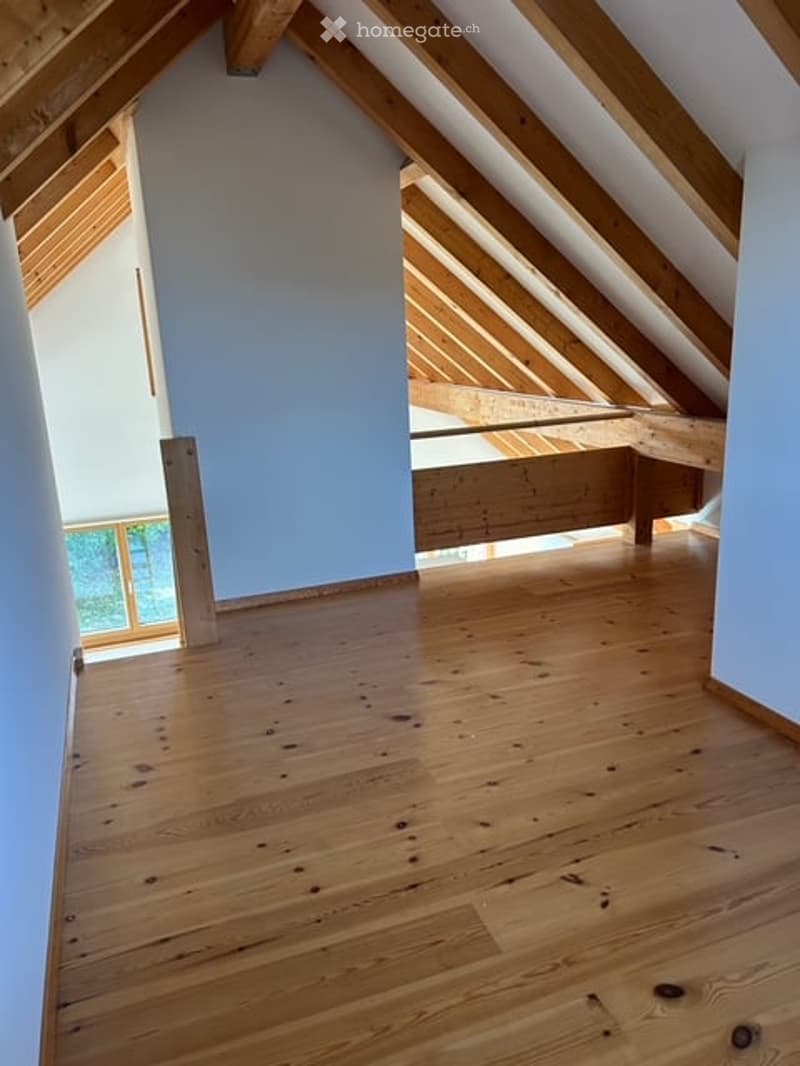 An attic space with exposed wooden beams, light blue walls, and a wooden floor. The room is completely empty with no furniture or equipment.