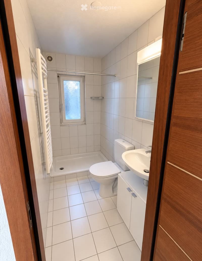 Modern bathroom with white tiles, a bathtub, toilet, sink, and a mirror. Features a window with frosted glass and a towel rack on the left side.