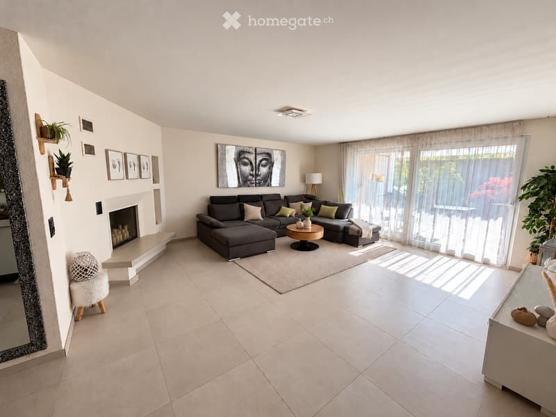 Modern living room with a grey sectional sofa, coffee table, fireplace, and large windows with sheer curtains. Features a white fur stool, decorative mirror, and several plants.