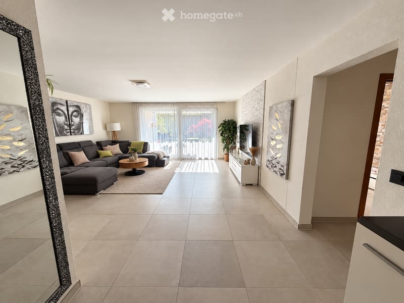 Modern living room with a grey sectional sofa, coffee table, side table, and artwork on the walls. The space features large windows with sheer curtains and a view of a balcony area.
