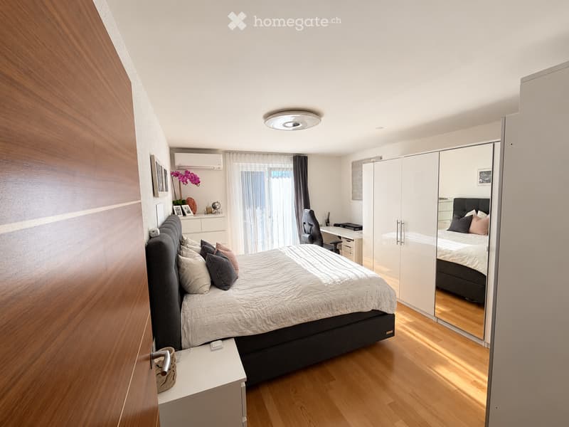 Modern bedroom with a large bed, nightstand, desk, chair, and large mirror wardrobe. The room features wooden flooring, large windows with curtains, and decorative items on the wall.