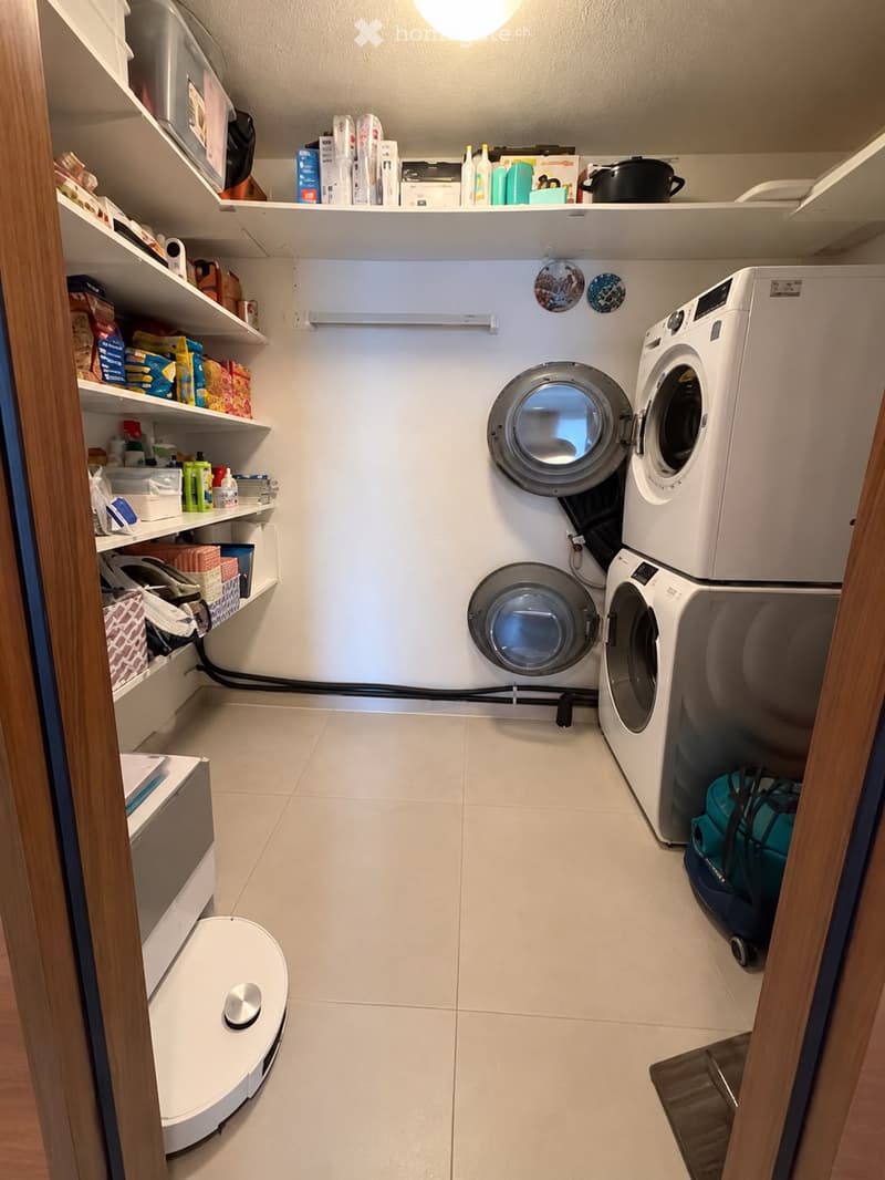 Well-organized laundry room with white tiled floor, white walls, and built-in shelves holding cleaning supplies and household items. Features a stacked washer and dryer unit, a robotic vacuum on the floor, and a small rug near the door.