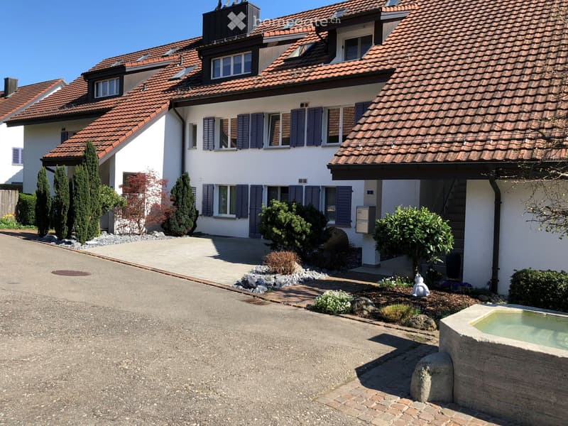 Two-story house with red-tiled roof, white walls, multiple windows with shutters, and a landscaped garden with a small pond and decorative elements.